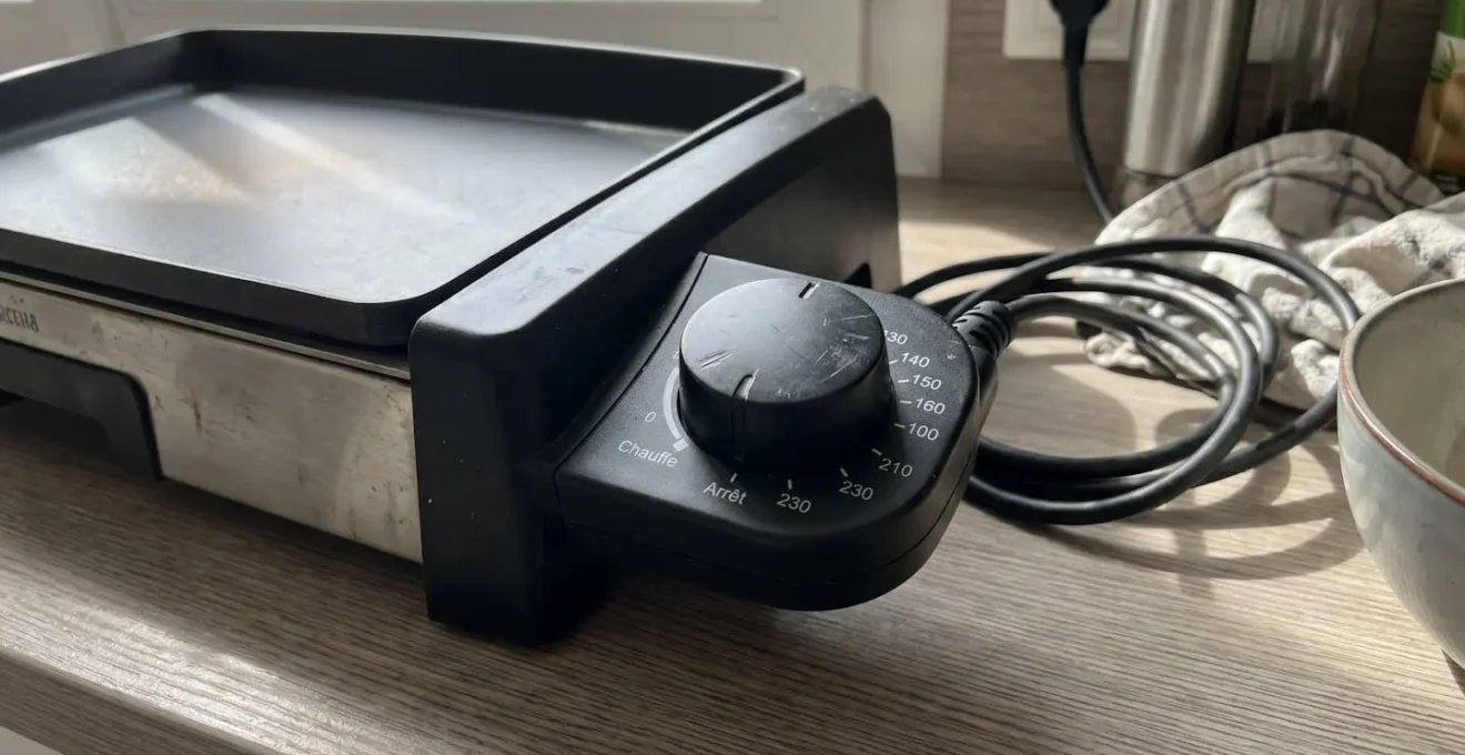 Gros plan sur le bouton de réglage du thermostat d'une plancha électrique avec graduations de température visibles et indicateur lumineux, posé sur un plan de travail en quartz blanc dans une cuisine contemporaine