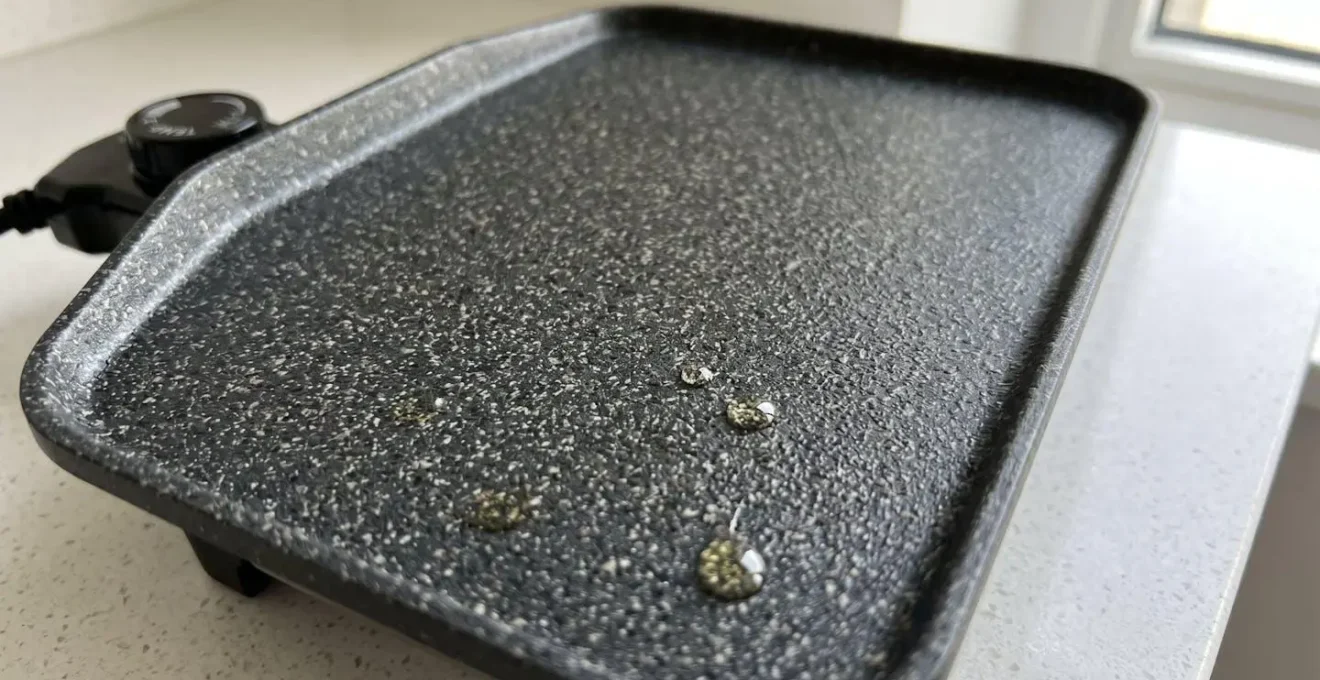 Gros plan macro sur la surface texturée d'un revêtement effet pierre d'une plancha électrique avec micro-motif granulaire et quelques fines gouttes d'huile d'olive reflétant la lumière naturelle